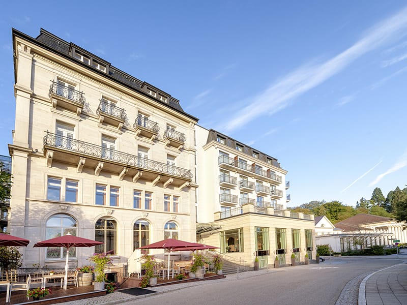 Außenansicht des Maison Messmer Luxushotel Baden Baden, mit heller Fassade, Terrasse und roten Sonnenschirmen