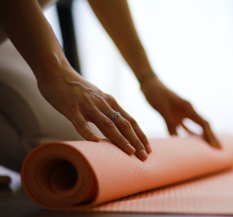 Nahaufnahme zweier Hände, wie sich eine orangene Yoga-Matte auf dem Boden ausrollen