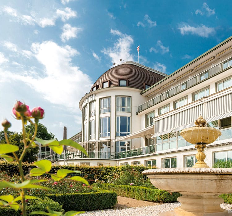 The exterior facade of the Parkhotel overlooking the garden with a fountain and flowers